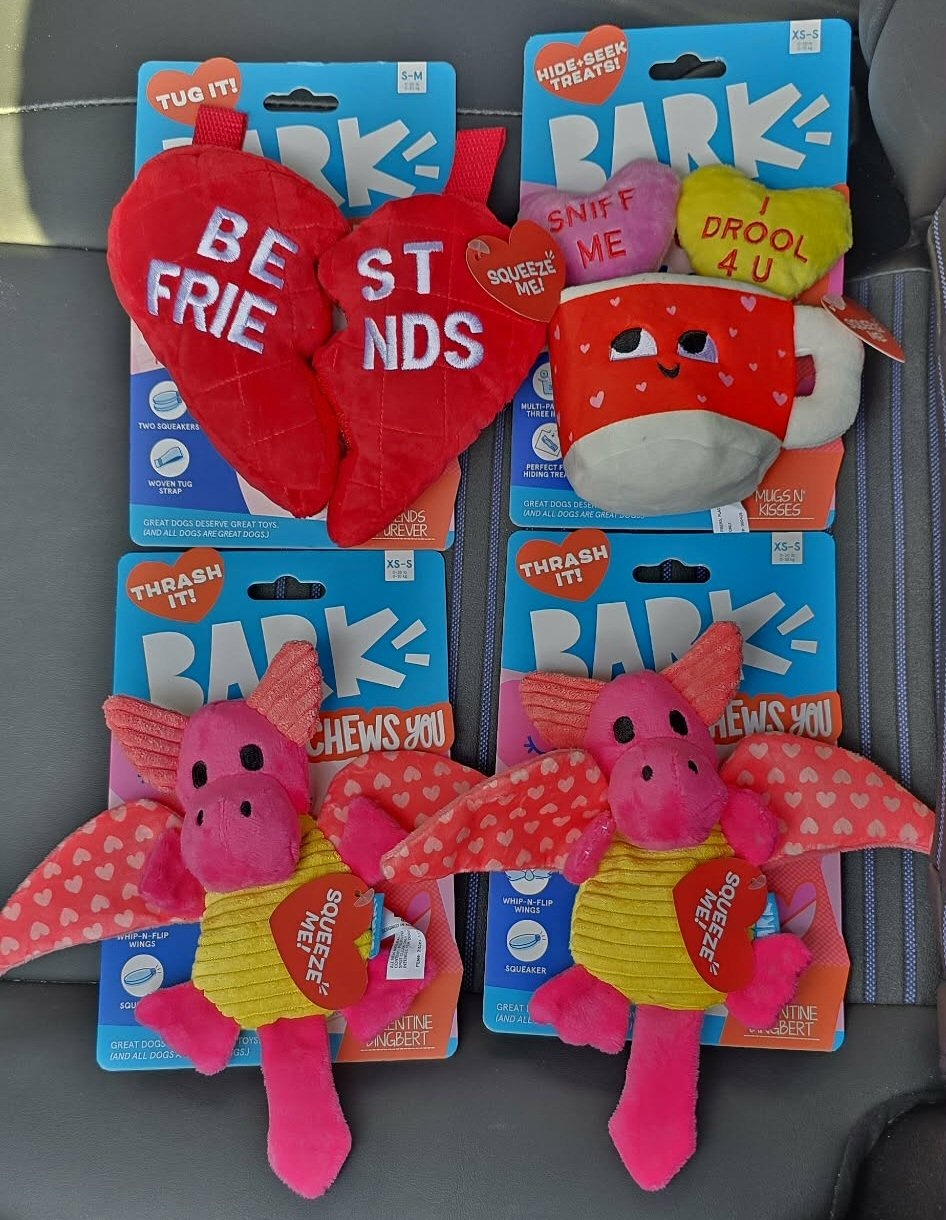 Four Bark brand Valentine-themed dog toys displayed on a car seat, including a red heart toy reading “best friends,” a plush mug toy with small hearts labeled “sniff me” and “i drool 4 u,” and two pink dragon toys with heart-pattern wings and squeeze me tags.