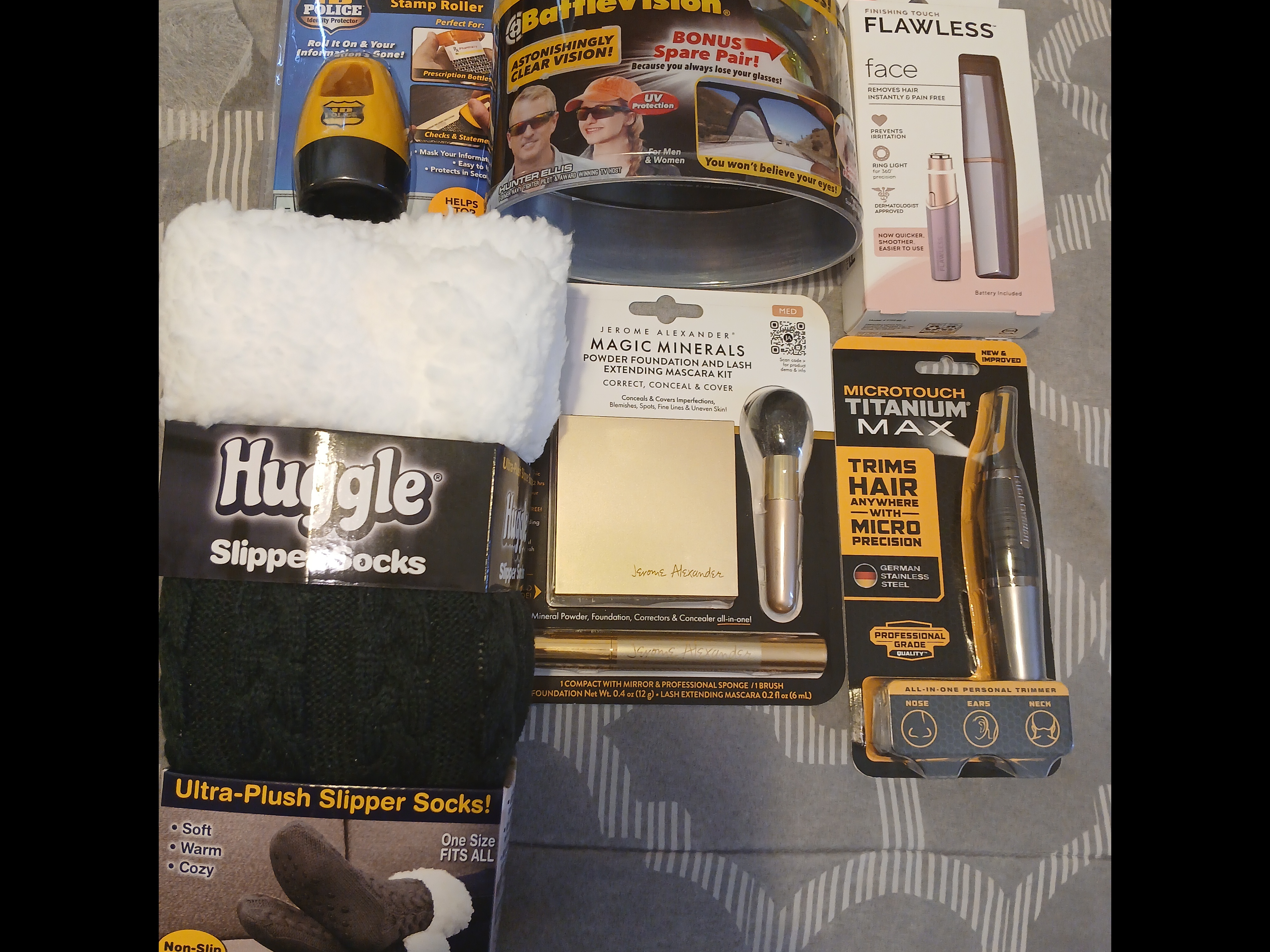 Assortment of retail products displayed on a surface, including Huggle ultra-plush slipper socks, BattleVision glasses with bonus spare pair, Finishing Touch Flawless facial hair remover, Jerome Alexander Magic Minerals powder foundation set with brush and mascara, MicroTouch Titanium Max precision trimmer, and a Guard Your ID roller.